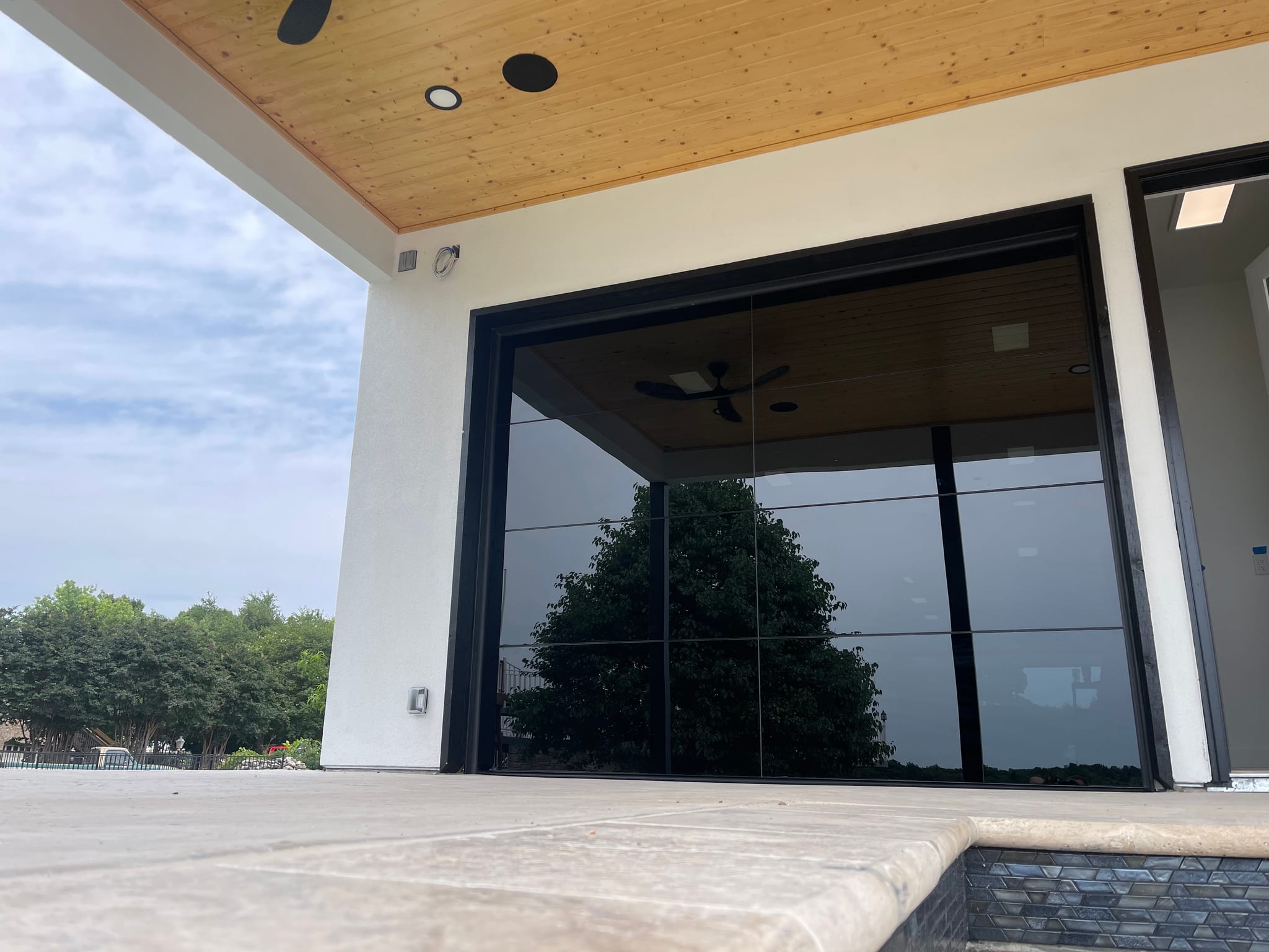 A black full view glass garage door in a white stucco wall beside a pool, reflecting a tree and the patio ceiling
