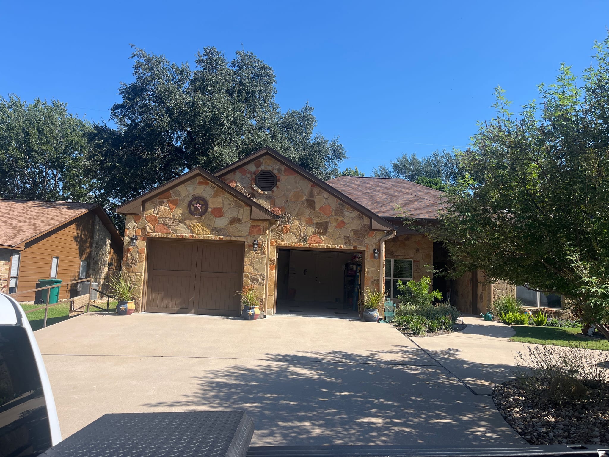 Two brown raised-panel garage doors on a stone-front home with a long concrete driveway and oak trees in Meadowlakes