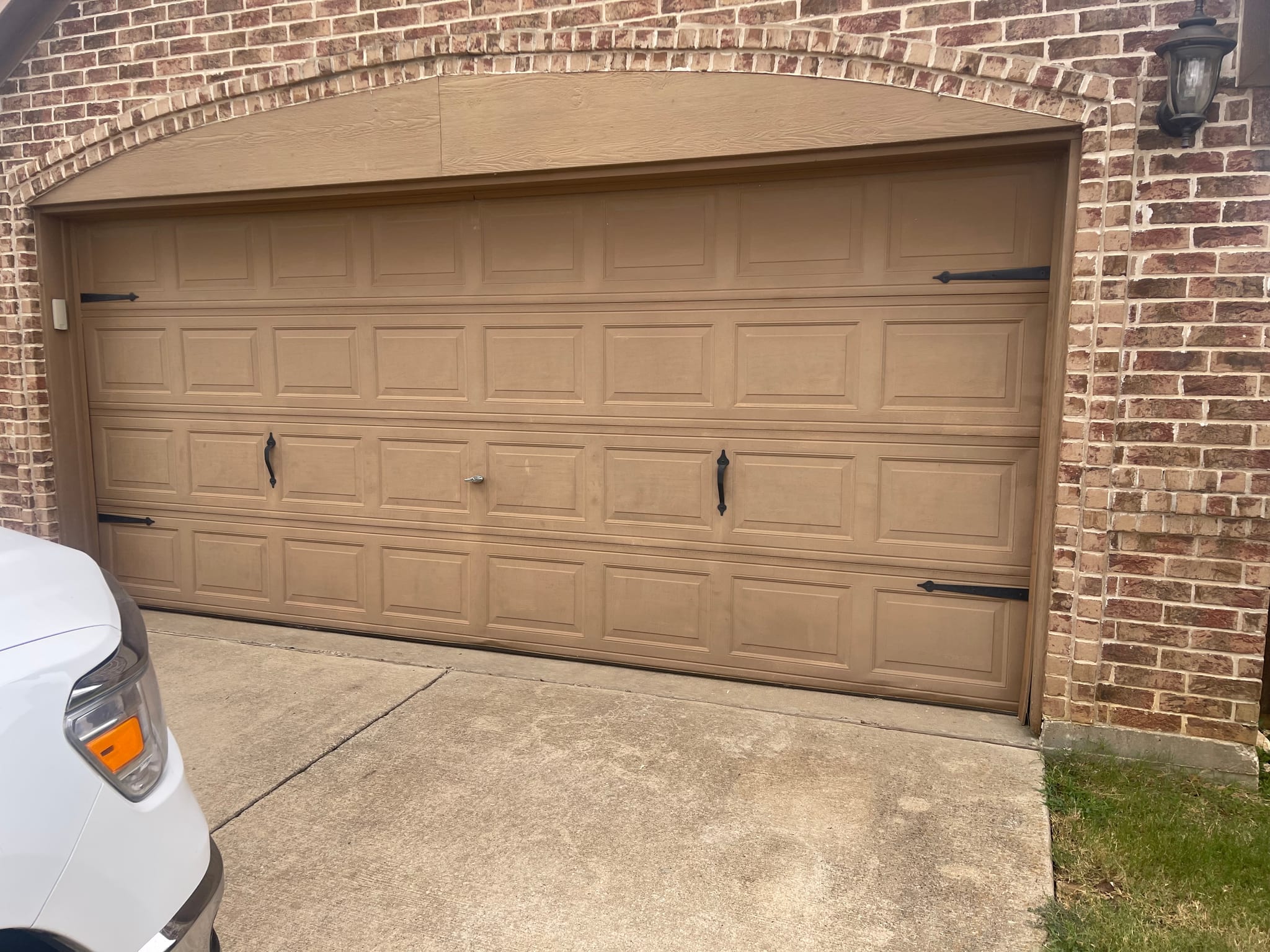 A tan double garage door with raised panels and decorative black hardware installed in a brick house